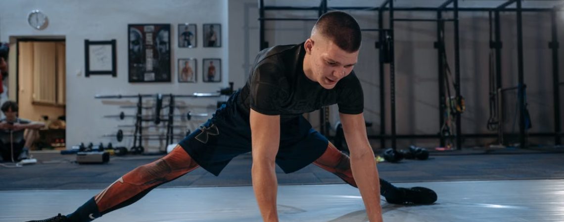 A male athlete stretches intensely in a dimly lit gym environment, preparing for training.